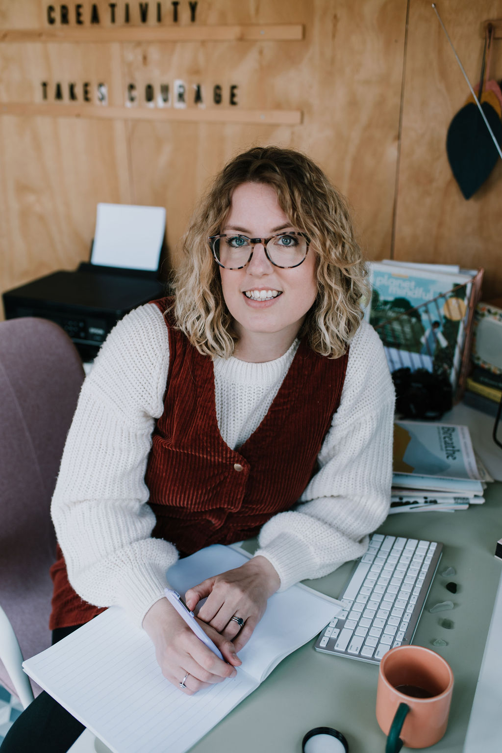 Clients of And So The Story Goes Ltd. Hayley sitting at her desk wearing a white jumper and orange dress leans towards the camera looking at the lens with a pen in her hand and a notebook. Hayley is proud of her CIPR certification.