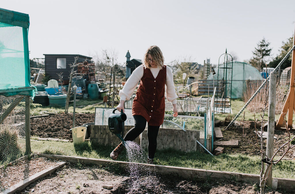Hayley is watering her allotment plot with a green watering can. Behind her is the other plots on the allotment