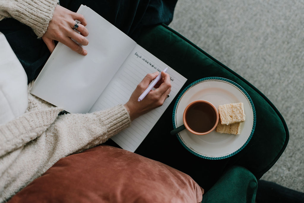 A notebook resting on the edge of a green sofa with a hand stretched across writing a blog entry. A cup of tea sits of a plate along with two small slices of cake