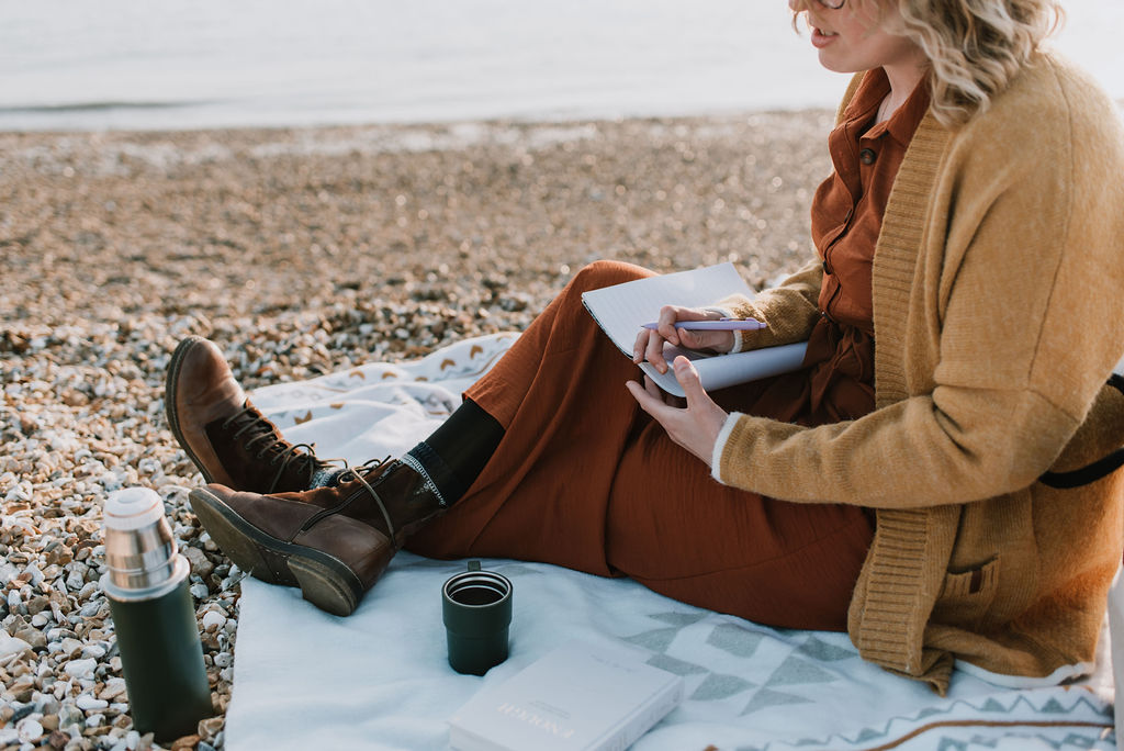 Hayley sitting on a blanket on a stoney beach holding a notebook and pen 