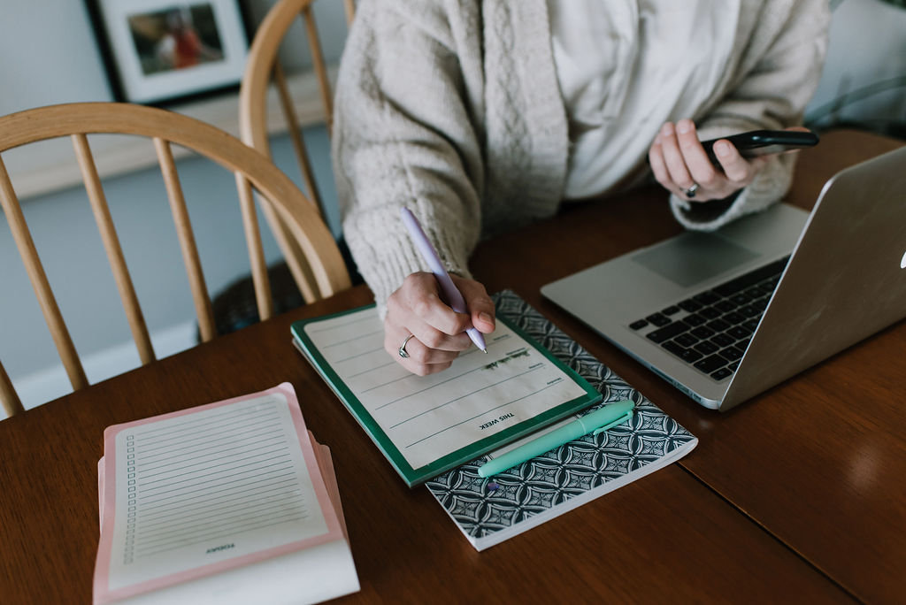 A close up of Hayley writing on a notepad. She's sitting at the table with her laptop in front of her holding her phone in her other hand.