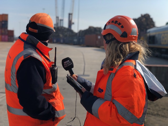 Woman in high vis holding a microphone between her and the male interviewee. both with their heads turned towards the rail track in the background