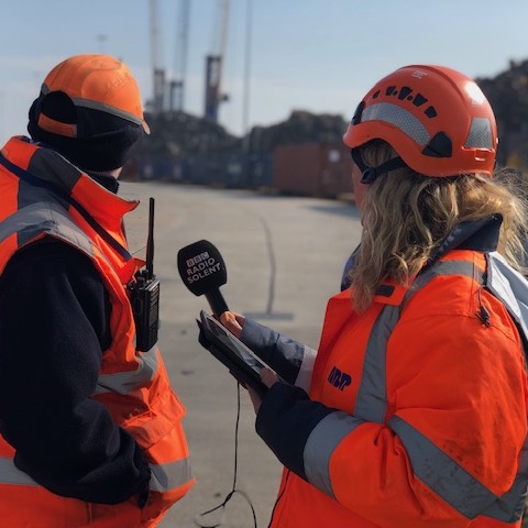 Maritime PR and communications - Woman in high vis holding a microphone between her and the male interviewee. both with their heads turned towards the rail track in the background